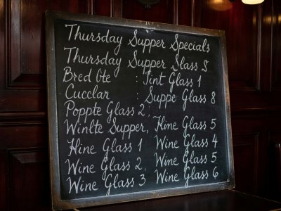 Handwritten blackboard menu leaning against a dark wainscoted wall showing Thursday supper specials and wine glass prices